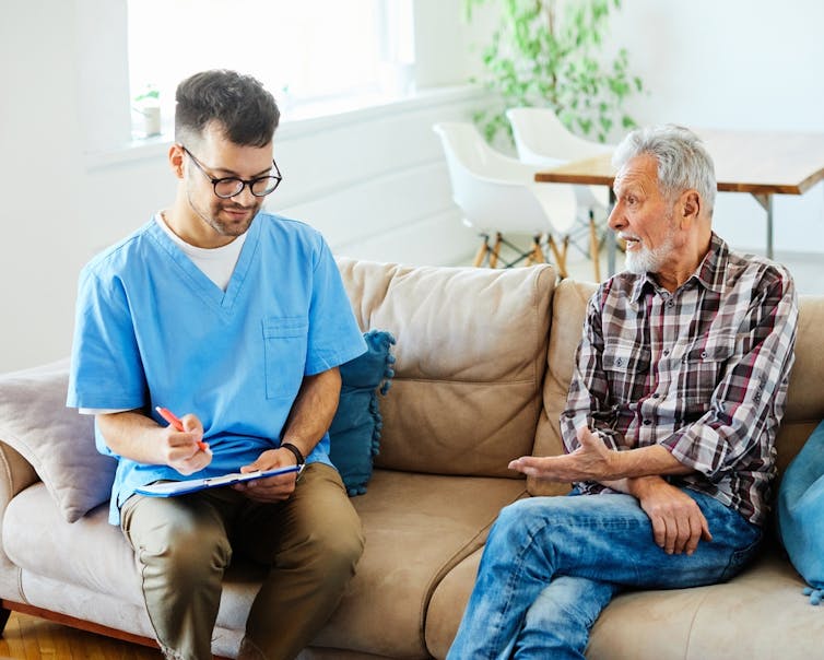 Male nurse and elderly male patient sitting on sofa, nurse writing on clipboard